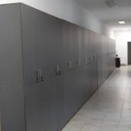 Długi rząd szarych szafek zamykanych na klucz, ciągnący się wzdłuż korytarza z kafelkową podłogą, narzędzia w torbie na pierwszym planie.