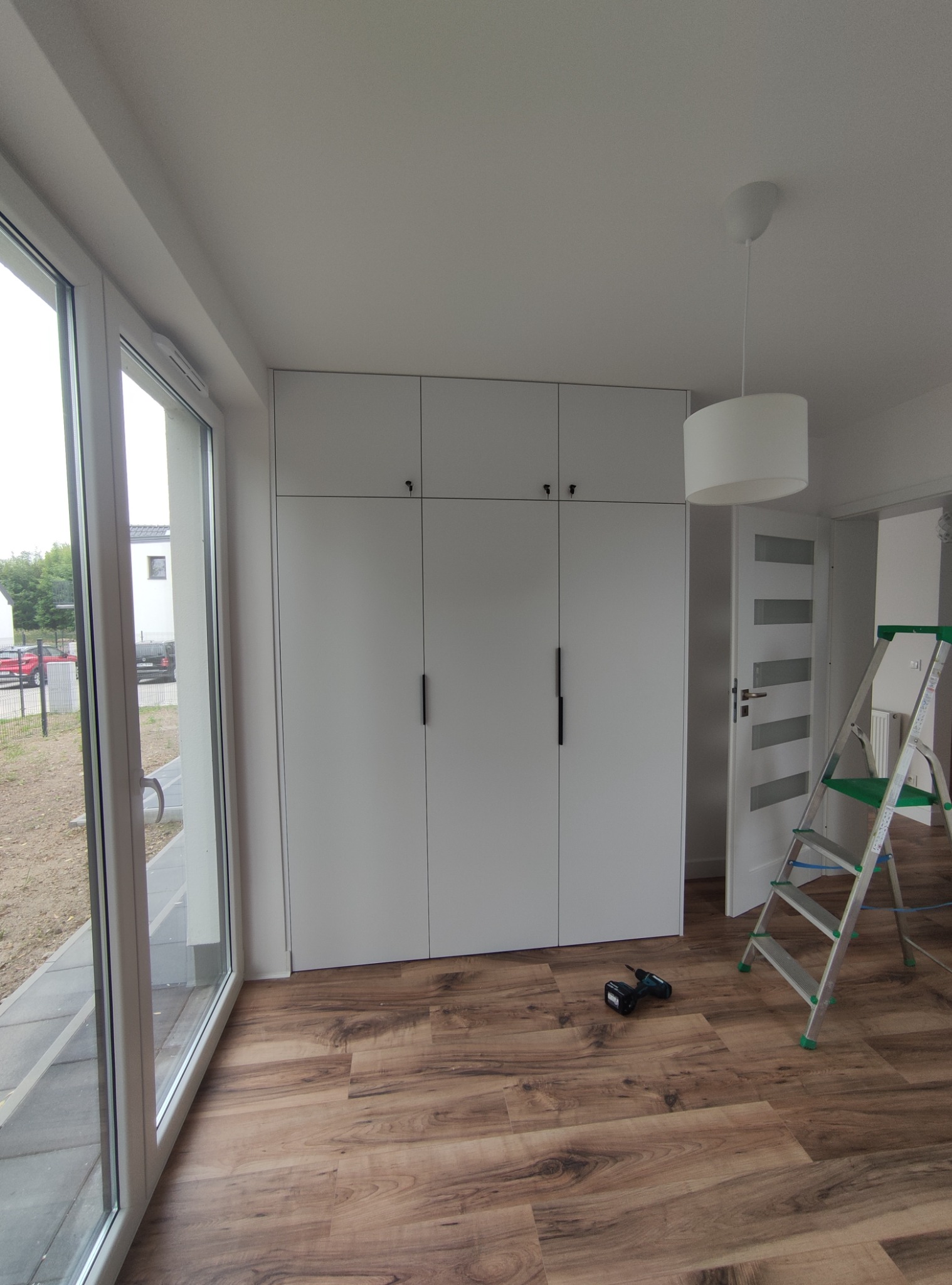 Biała szafa na wymiar z długimi uchwytami w jasnym pokoju, widoczna wiertarka i drabina, parkiet w odcieniach brązu.