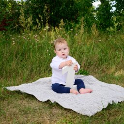 Katarzyna Olkowska Photo Memory - Roczne dziecko w białej koszuli i granatowych spodniach siedzi na szarym kocu w trawie, trzymając białą cyfrę '1'.