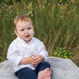 Katarzyna Olkowska Photo Memory - Mały chłopiec w białej koszuli i granatowych spodniach siedzi boso na szarym kocu na tle wysokiej trawy z małymi różowymi kwiatkami, z opatrunkiem na policzku.