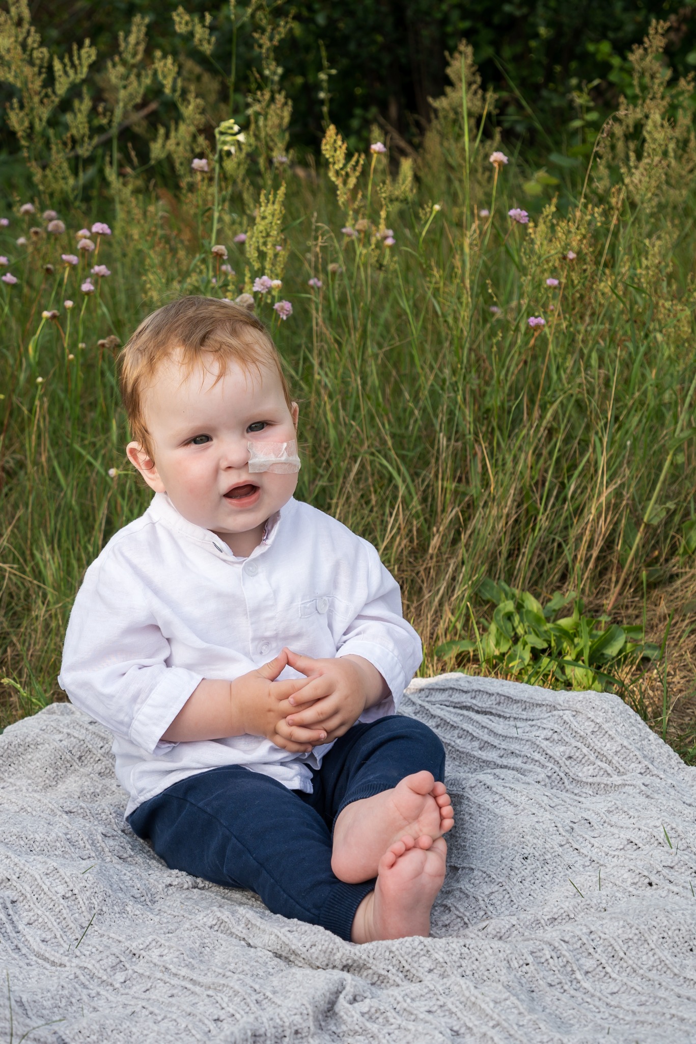 Mały chłopiec w białej koszuli i granatowych spodniach siedzi boso na szarym kocu na tle wysokiej trawy z małymi różowymi kwiatkami, z opatrunkiem na policzku.