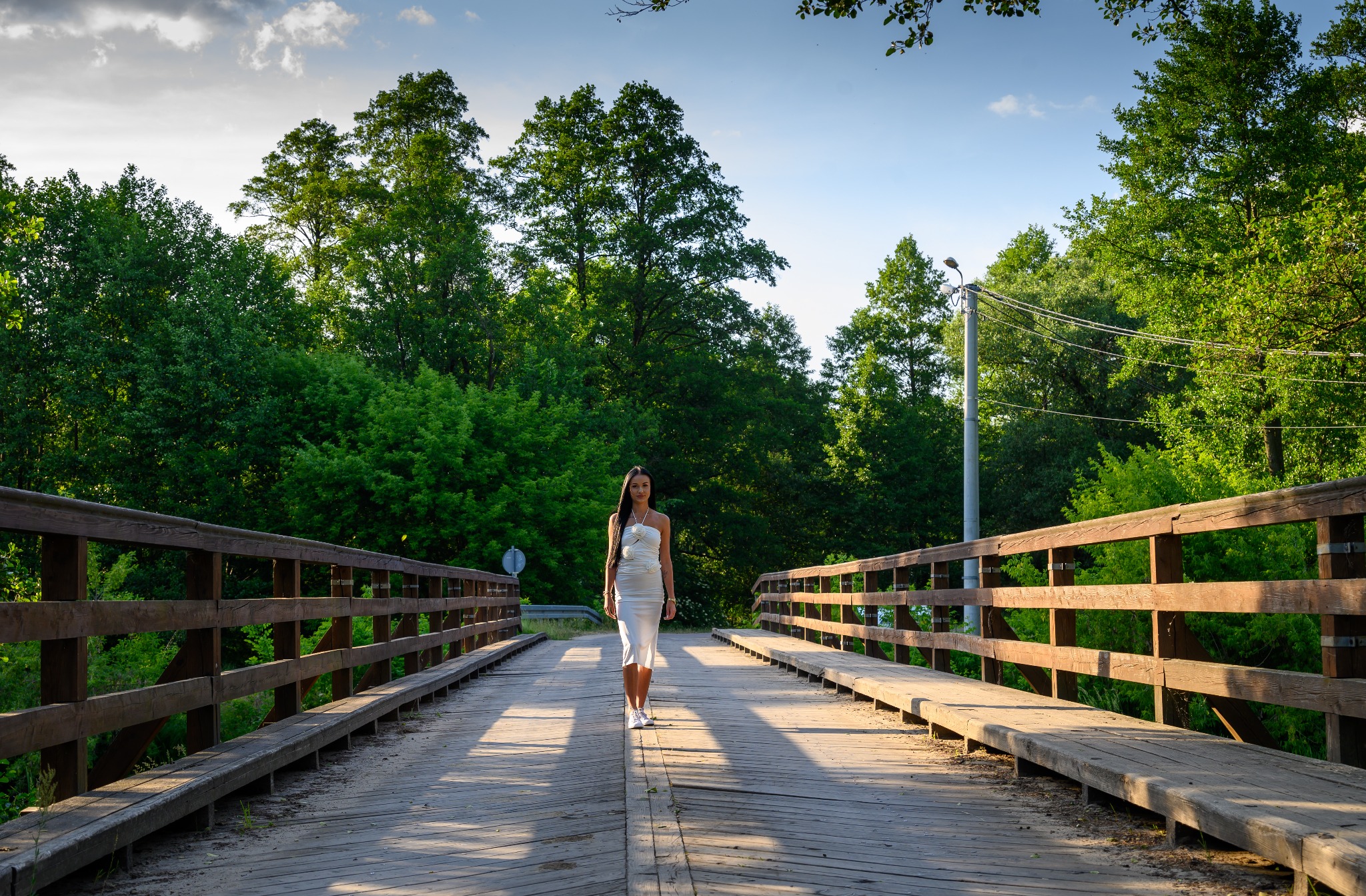Kobieta w białej sukience spaceruje po drewnianym moście na tle zieleni drzew i błękitnego nieba.