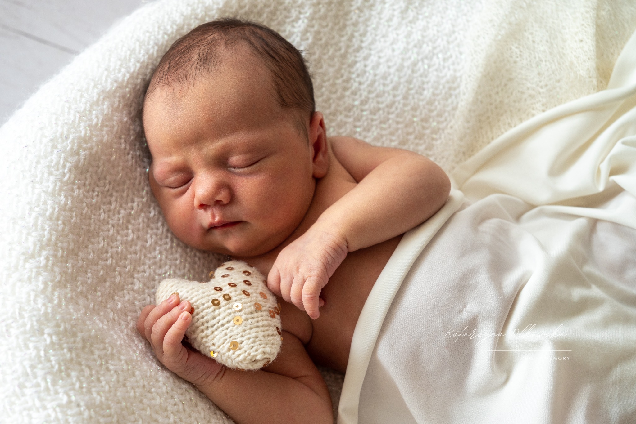 Śpiący noworodek otulony białymi tkaninami, trzymający w dłoni dziergane serduszko z cekinami.