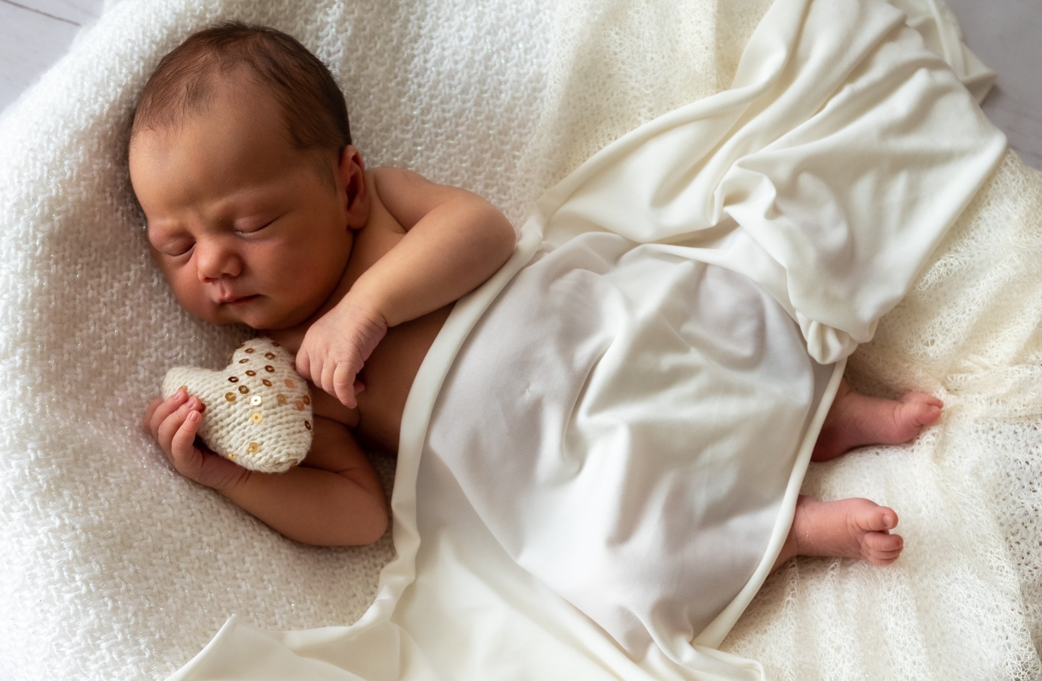 Noworodek śpiący na puszystym kocu, otulony kremowym materiałem, trzymając w dłoni dziergane serce z cekinami.