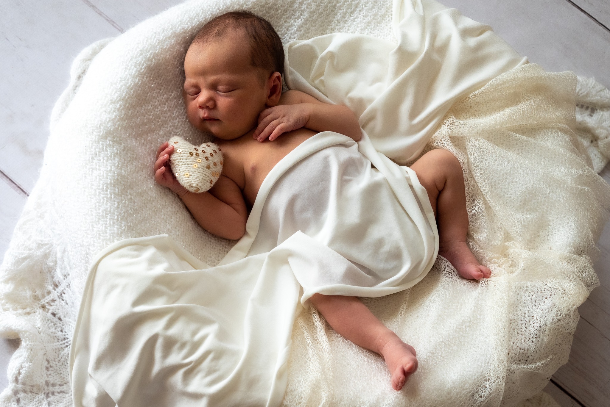 Śpiący noworodek owinięty w białe tkaniny, trzymający w dłoni małe serce z cekinami, leżący na puszystym, białym kocyku.