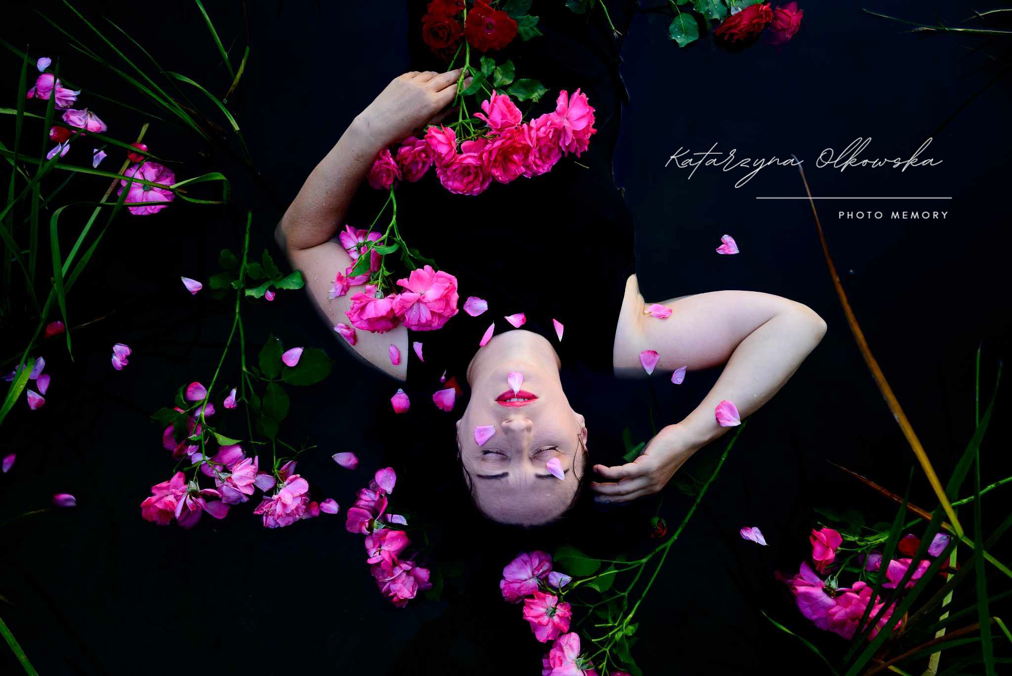 Kobieta leżąca w ciemnej wodzie otoczona różowymi i czerwonymi różami, płatki róż na twarzy i wodzie, artystyczna sesja w stylu dark floral.