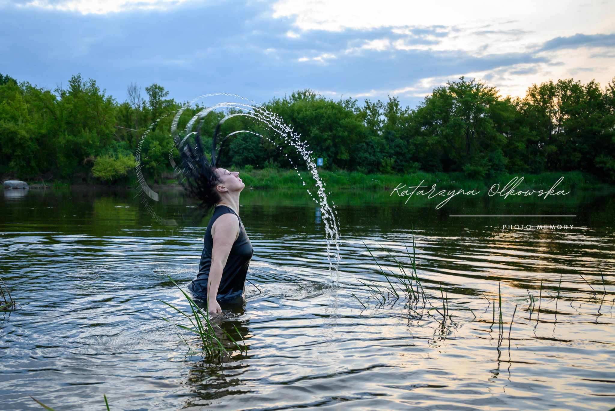 Kobieta w czarnym stroju odrzuca mokre włosy w rzece, tworząc efektowny wachlarz kropli wody, tło stanowią drzewa i pochmurne niebo o zmierzchu, w rogu widoczny podpis fotografa.