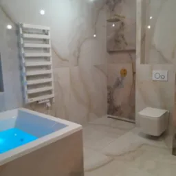 Elegancka łazienka wykończona płytkami imitującymi marmur, wanna z podświetleniem LED, grzejnik drabinkowy, bateria prysznicowa w kolorze złotym i podwieszana toaleta.