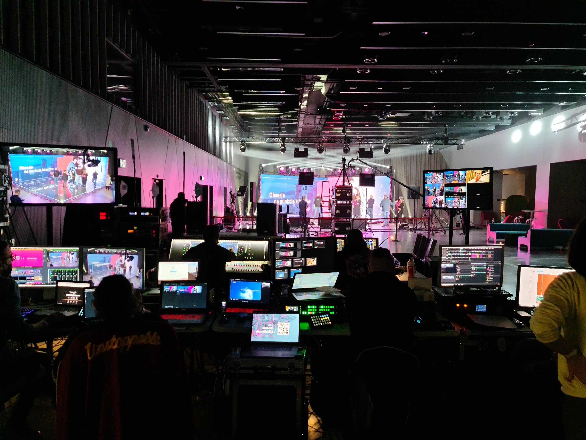 Reżyserka podczas nagrywania programu telewizyjnego: monitory podglądowe, mikser wizyjny, komputery i ekipa techniczna w zaciemnionym pomieszczeniu z widokiem na oświetloną scenę.