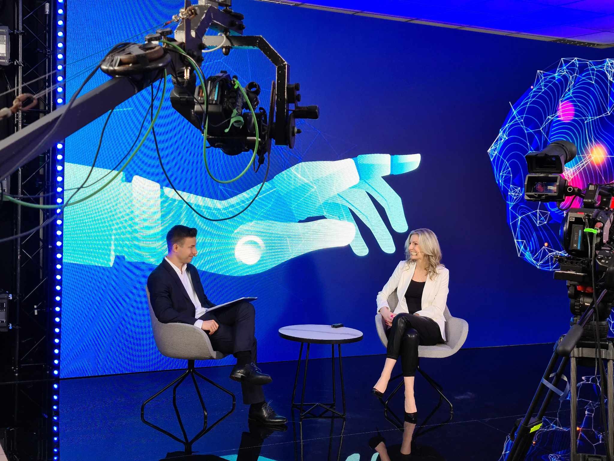 Studio nagraniowe z kamerą na wysięgniku, mężczyzną z dokumentami i kobietą w białej marynarce podczas wywiadu na tle cyfrowej grafiki.