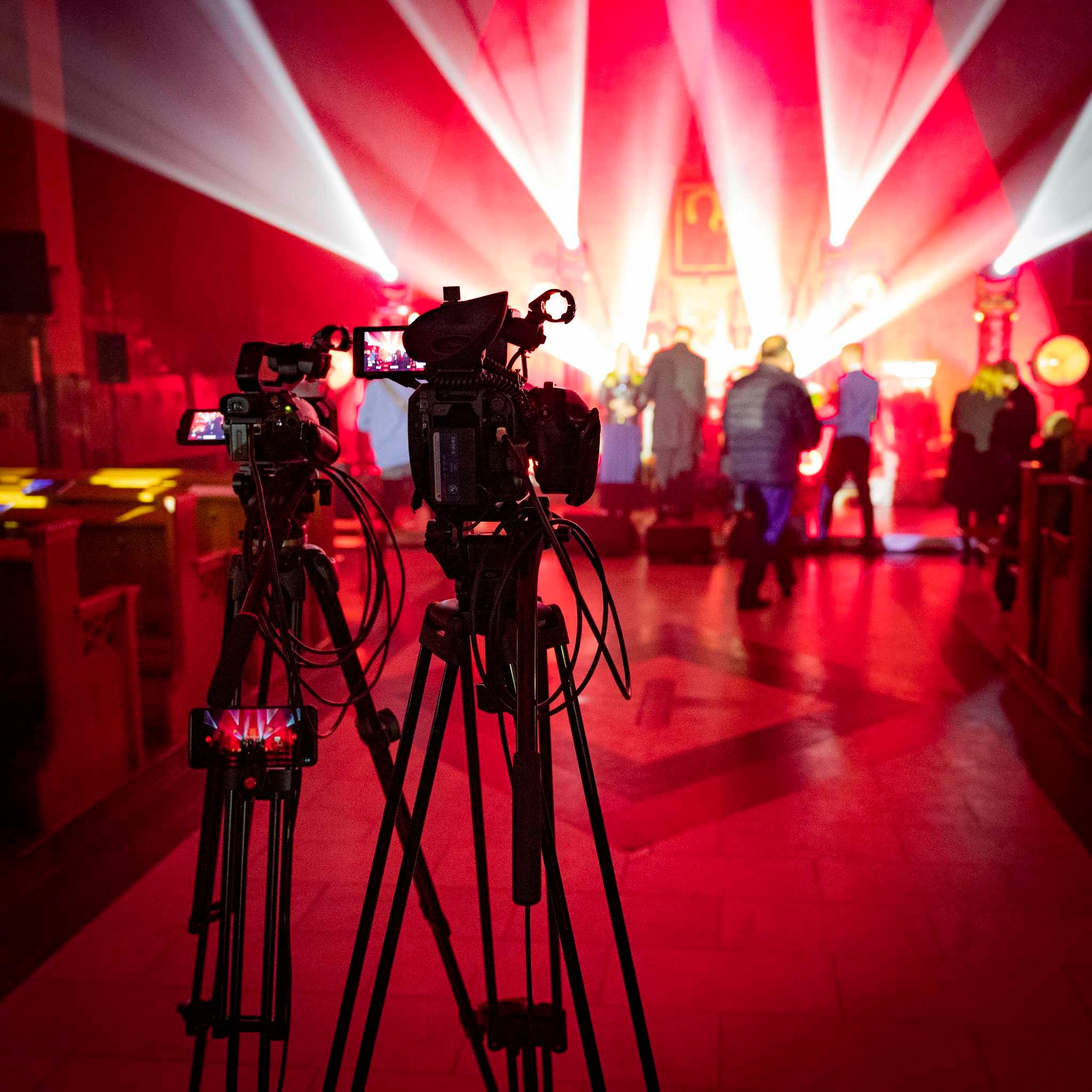 Dwa kamery wideo na statywach ustawione przed sceną oświetloną czerwonymi reflektorami podczas wydarzenia.