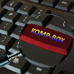 Klawiatura komputera z powiększoną lupą na klawiszu z napisem 'KOMP-BOX' w kolorach czerwonym i niebieskim, prawdopodobnie nazwa firmy lub produktu.