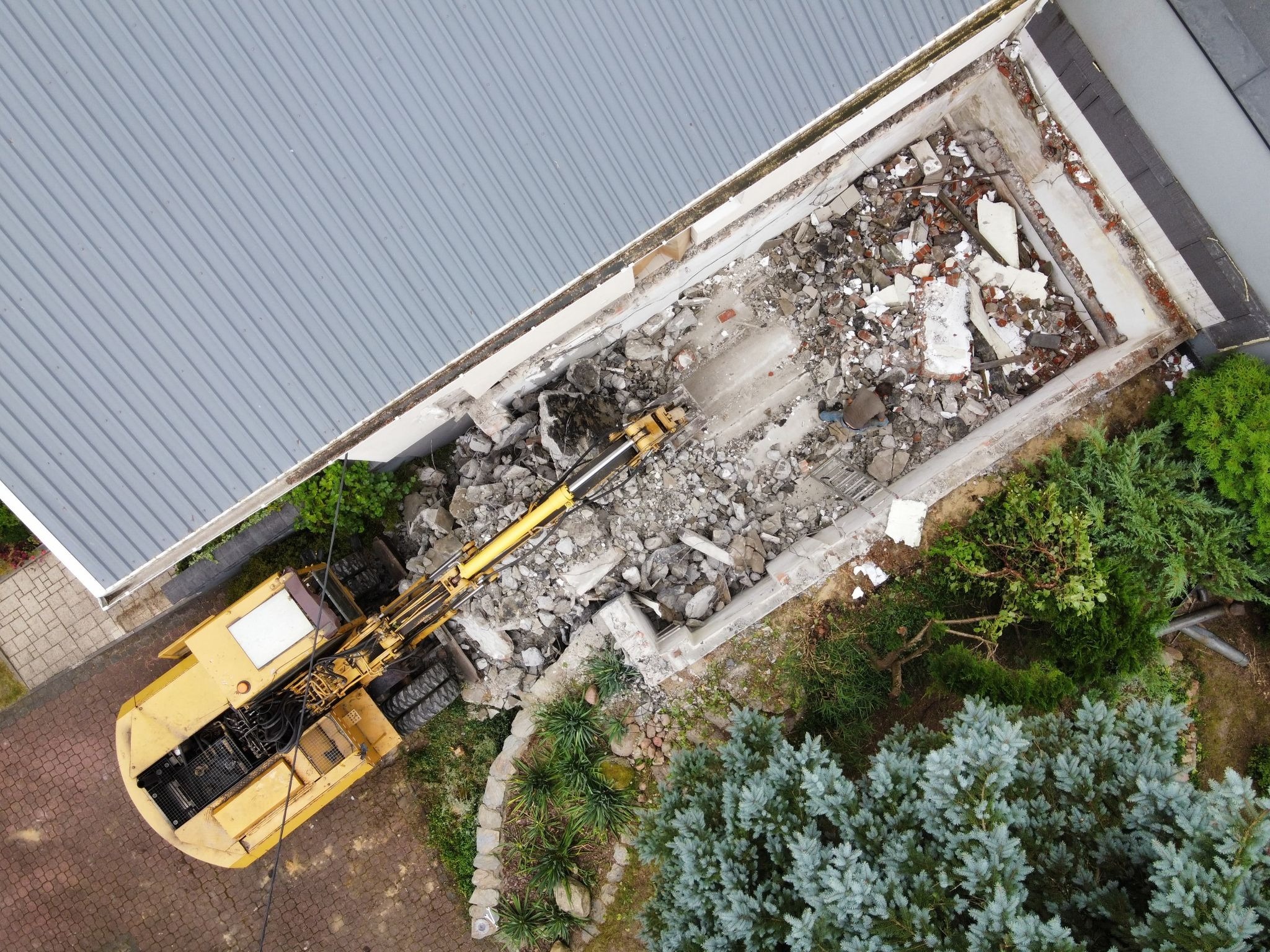 Widok z góry na wyburzanie przybudówki domu jednorodzinnego za pomocą koparki, gruz wewnątrz fundamentów, obok rosną iglaki.