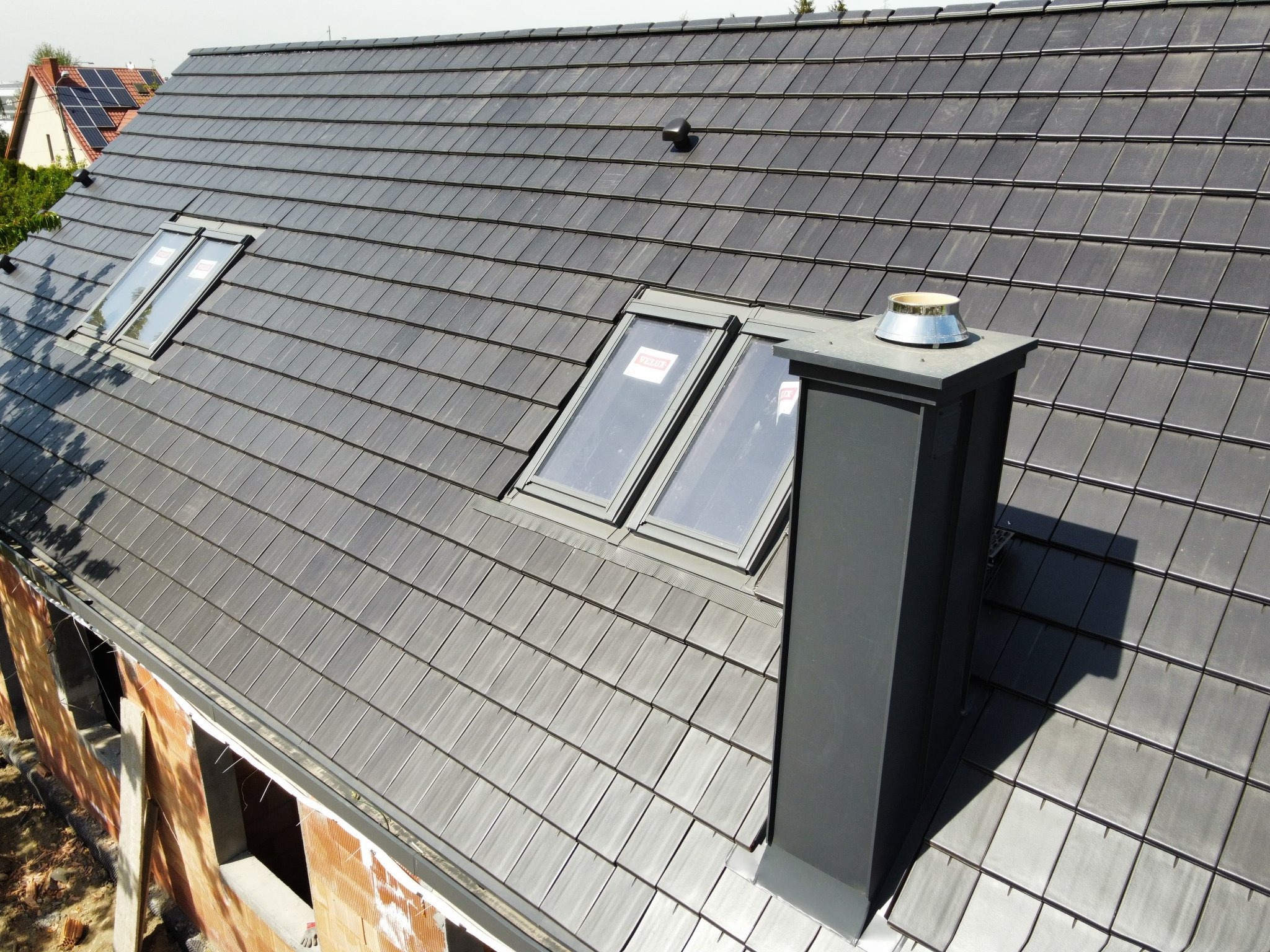 Nowy dach z ciemnej dachówki ceramicznej z dwoma oknami dachowymi i kominem, widok z góry na budynek w trakcie budowy.