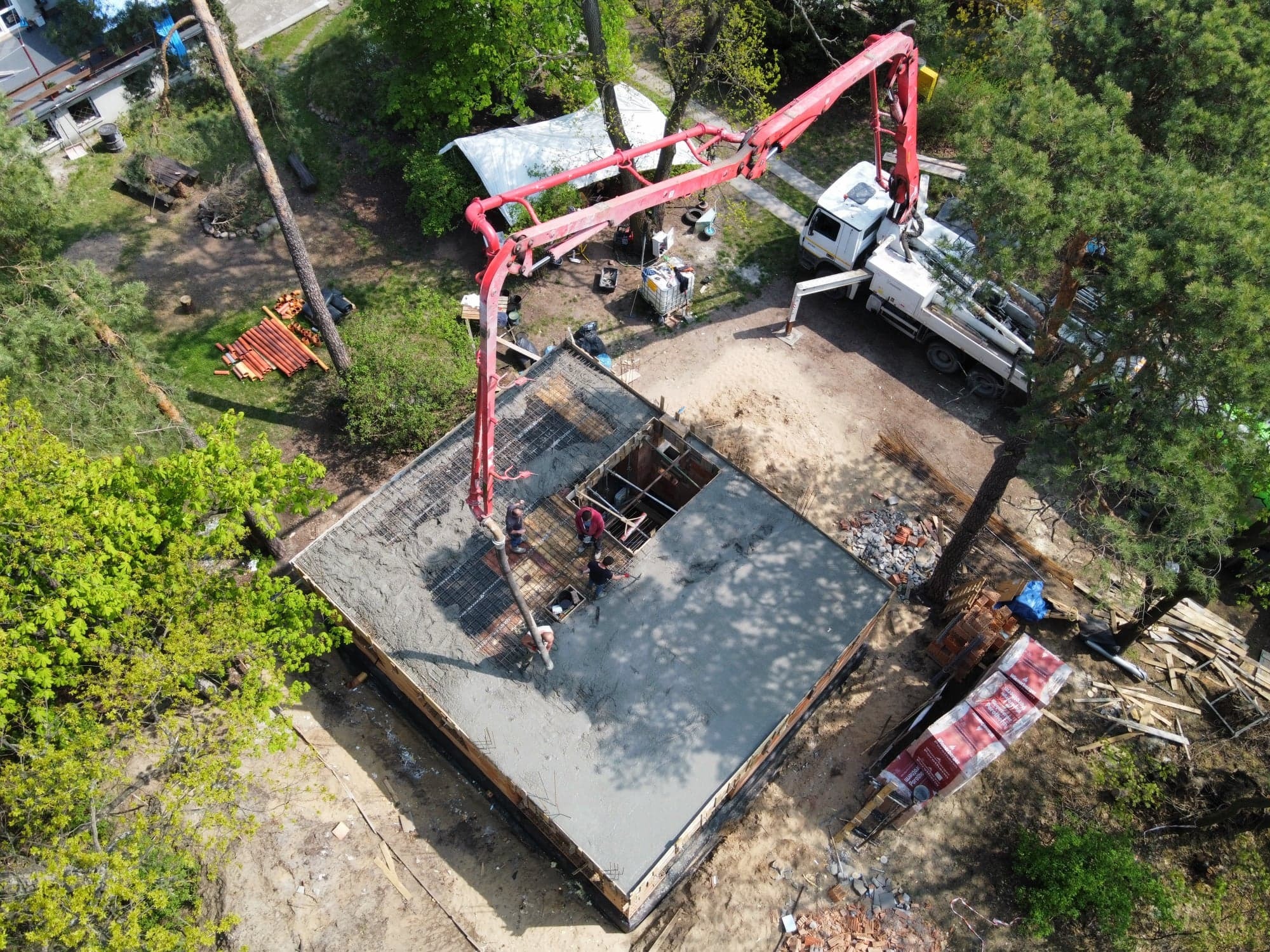 Zalewanie betonem fundamentów domu jednorodzinnego z użyciem pompy do betonu, widok z drona na plac budowy otoczony zielenią.
