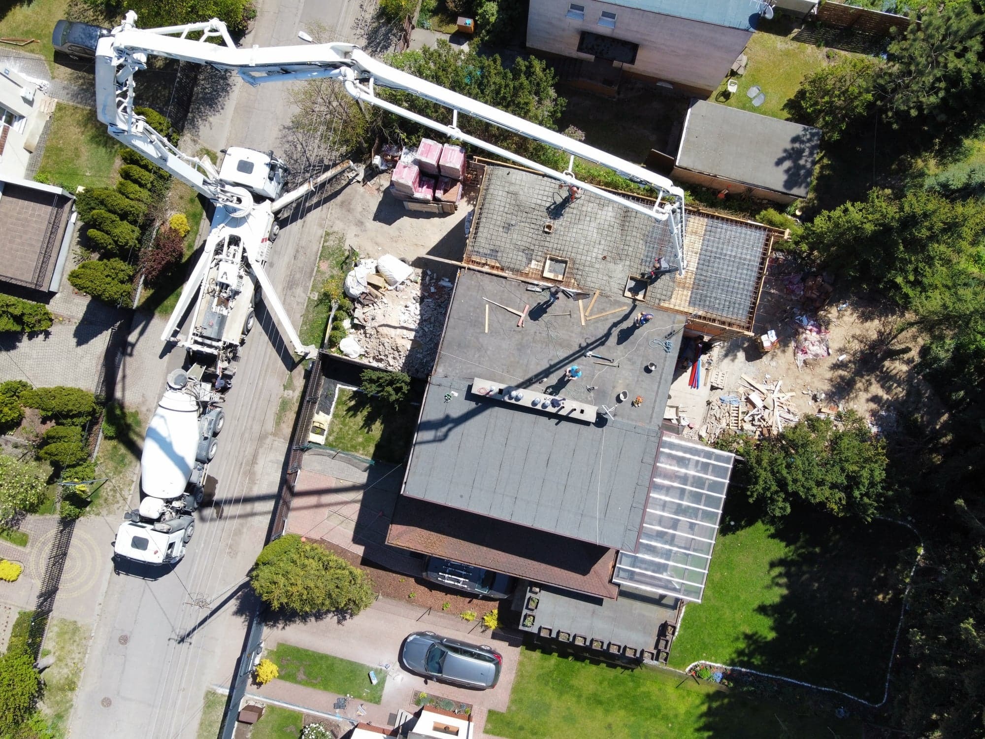 Widok z drona na budowę domu: betonowanie stropu z użyciem pompy do betonu, widoczni pracownicy, zbrojenie, palety z cegłami, gruz i częściowo wykończony budynek obok.