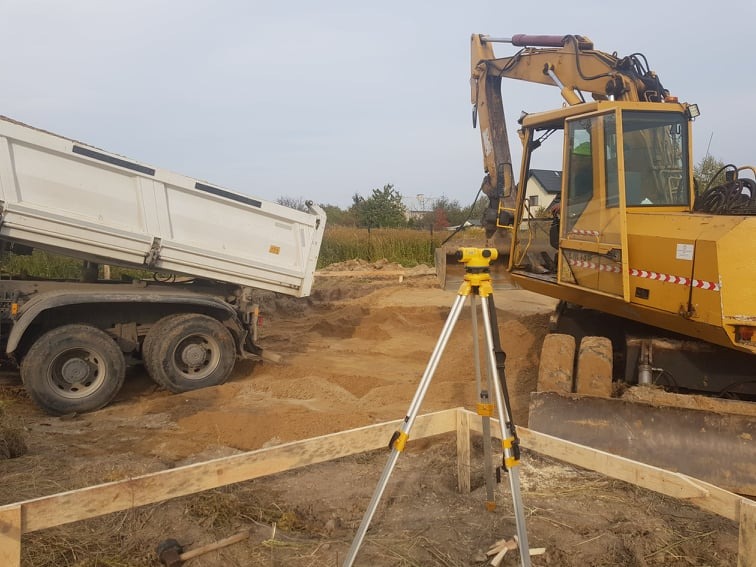 Przygotowanie terenu pod budowę: wywrotka wysypuje piasek, obok stoi żółta koparka, a w pierwszym planie niwelator na statywie i drewniane szalunki.