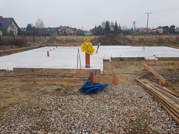 Wylana płyta fundamentowa pod dom murowany, z widocznymi pionowymi rurami instalacyjnymi zabezpieczonymi żółtą folią, deskowaniem i niebieskimi rurami instalacji wodnej na żwirowym podłożu.