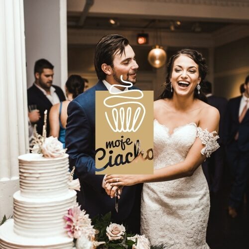 Para młoda w Suwałkach kroi tort weselny, zasłonięta częściowo kartką z napisem 'moje ciacho' i rysunkiem babeczki.