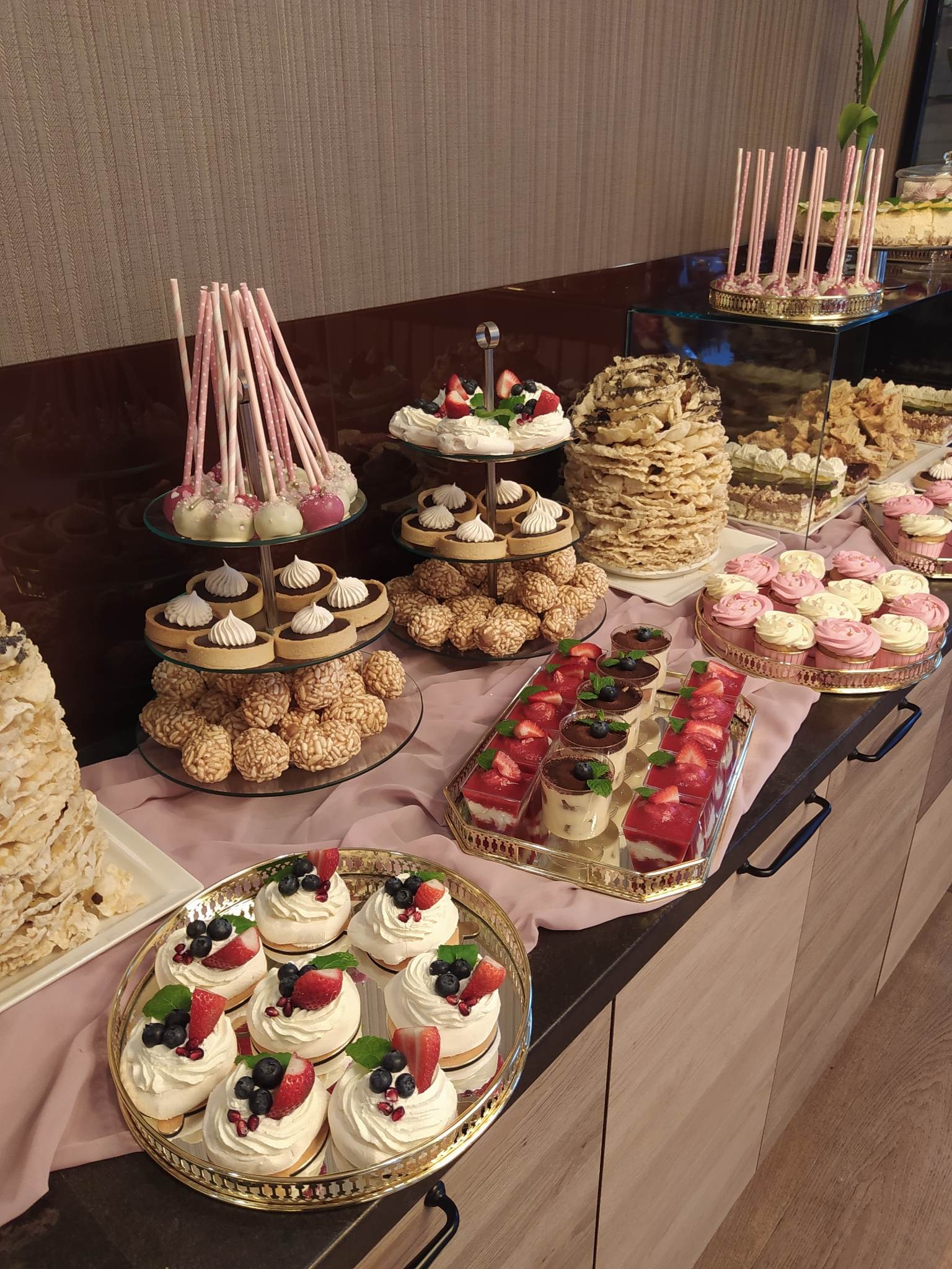 Bogato zastawiony stół z różnorodnymi deserami: babeczki z kremem, mini bezy z owocami, desery w szklankach z truskawkami, lizaki na patyku, torty bezowe, ciastka z kremem, wszystko na złotych...