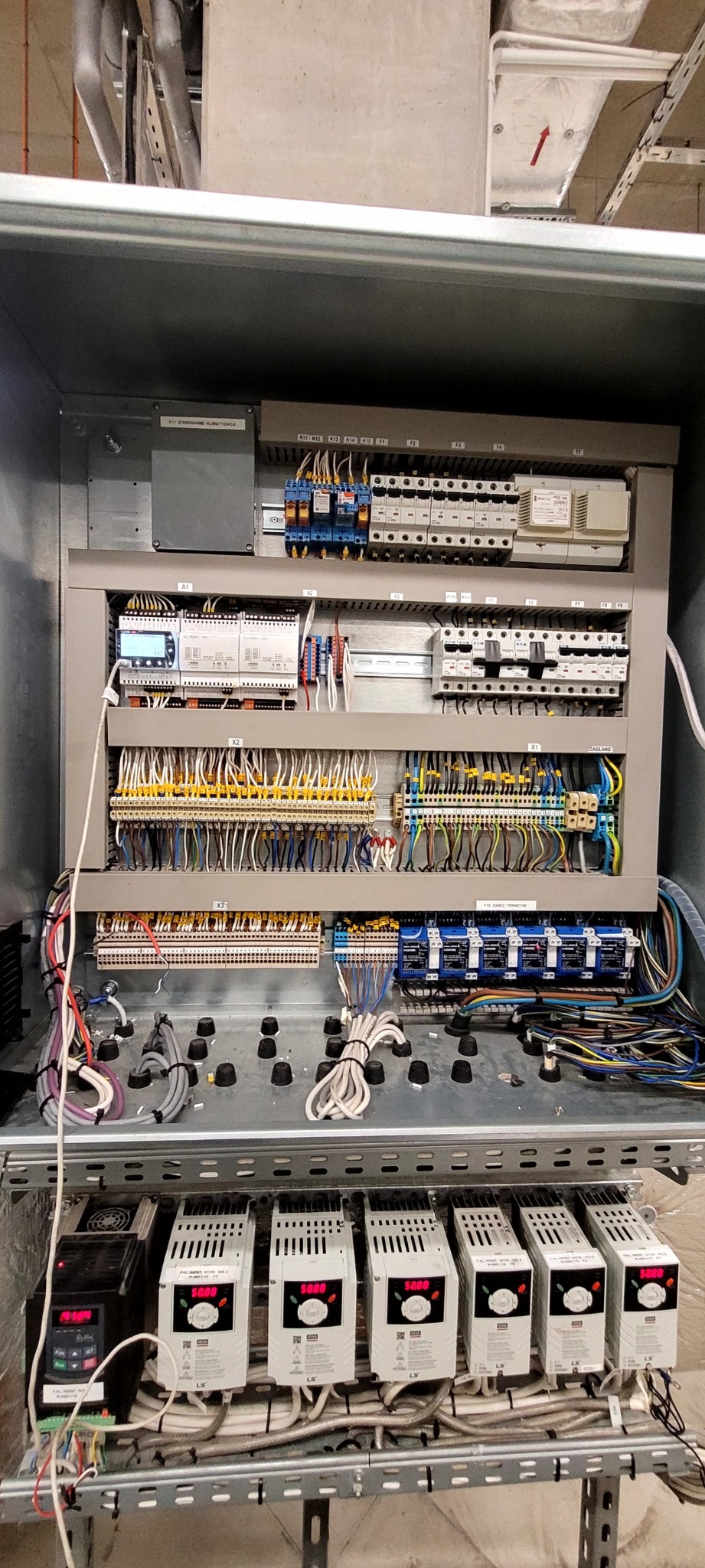 Panel sterowania klimatyzacją w metalowej obudowie, widoczne liczne przewody, sterowniki, bezpieczniki, wyświetlacze oraz moduły LS, całość zamontowana na metalowej konstrukcji w pomieszczeniu...