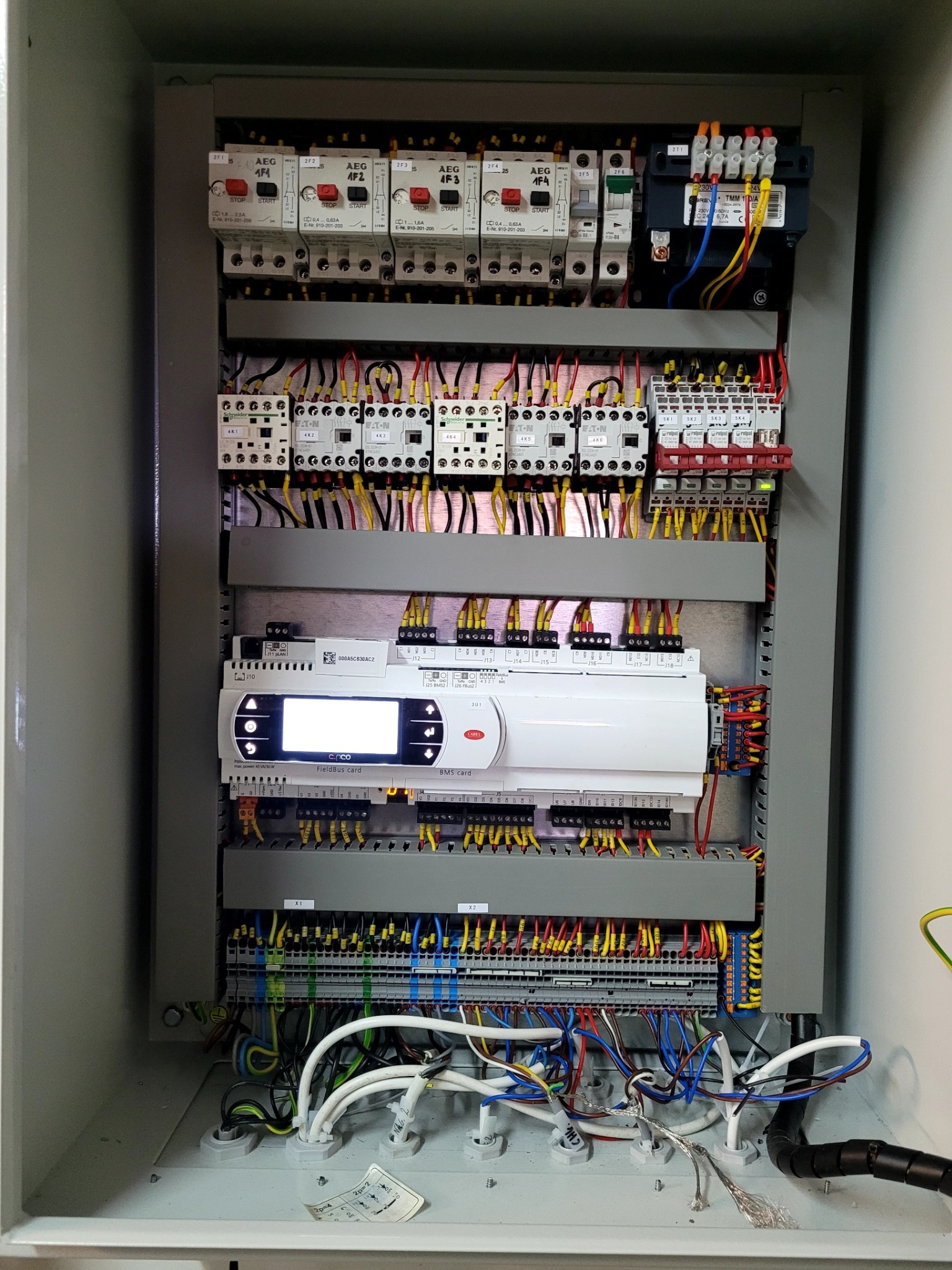 Wnętrze szarej szafy sterowniczej z widocznymi elementami elektrycznymi, w tym przekaźnikami AEG, Schneider Electric, Eaton, modułem sterującym c.pco FieldBus card i licznymi przewodami o różnych...