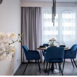 Jadalnia w nowoczesnym stylu: okrągły stół z drewnianym blatem, obity aksamitem krzesła w kolorze głębokiego błękitu, minimalistyczna lampa wisząca, białe storczyki w wazonie, szare zasłony.