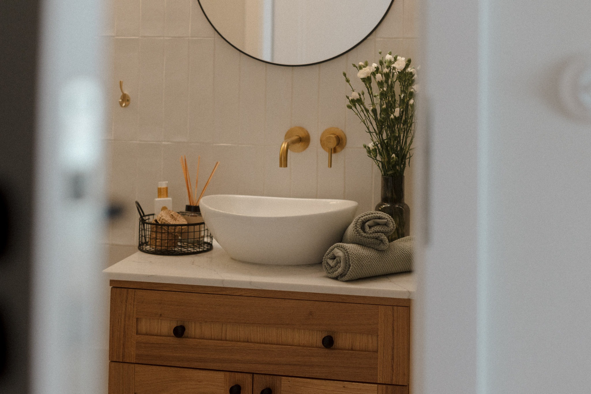 Nowoczesna łazienka z drewnianą szafką pod umywalką, marmurowym blatem, okrągłym lustrem i złotą armaturą, dekorowana kwiatami w wazonie i zwiniętymi ręcznikami.