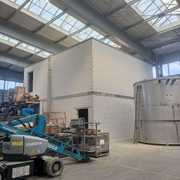 lintar group - Wnętrze hali produkcyjnej z wznoszoną dwukondygnacyjną konstrukcją z białych bloczków, widoczny podnośnik koszowy i zbiornik ze stali nierdzewnej, oświetlenie górne przez świetliki w dachu.