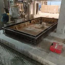 Budowa fundamentu: szalunek zbrojony stalą, przygotowany do zalania betonem, widoczna czerwona skrzynka narzędziowa Hilti, w tle maszyny i materiały budowlane.