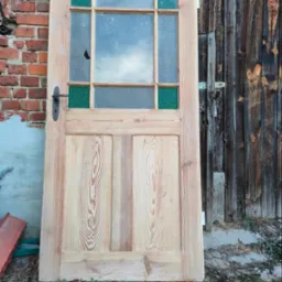 Odrestaurowane drewniane drzwi z szybami, z zielonymi akcentami, oparte o ceglany mur i drewnianą ścianę. Widoczne ślady renowacji i charakterystyczne usłojenie drewna.