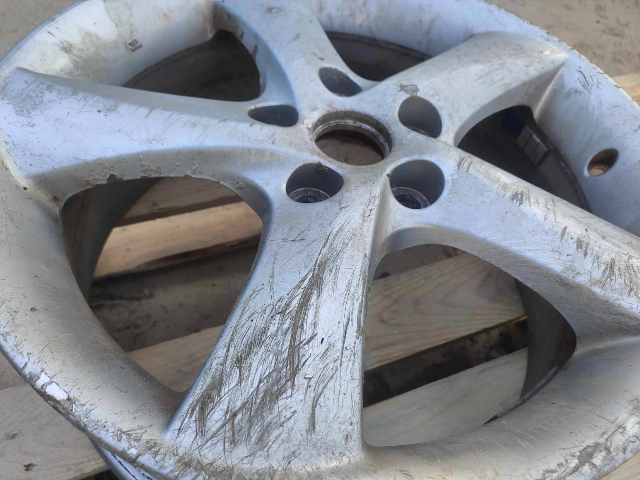 Brudna, aluminiowa felga samochodowa z widocznymi śladami zarysowań i zabrudzeń, leżąca na drewnianej palecie.