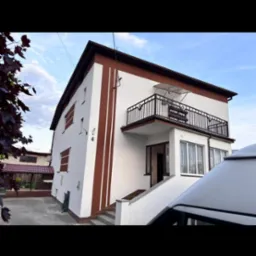 Dwupiętrowy dom z balkonem, białą elewacją i brązowymi pasami dekoracyjnymi. Widoczne schody wejściowe i część zaparkowanego pojazdu z przodu.