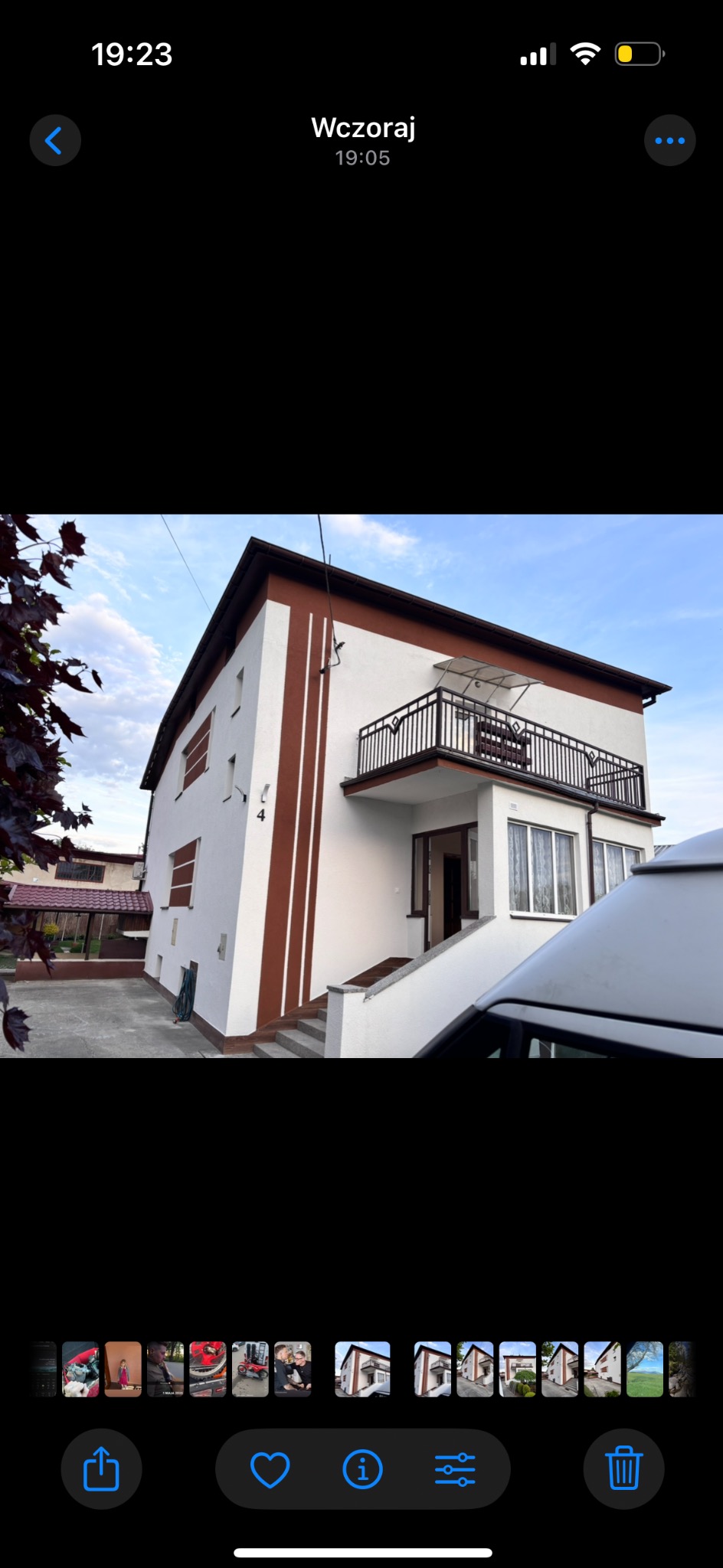 Dwupiętrowy dom z balkonem, białą elewacją i brązowymi pasami dekoracyjnymi. Widoczne schody wejściowe i część zaparkowanego pojazdu z przodu.