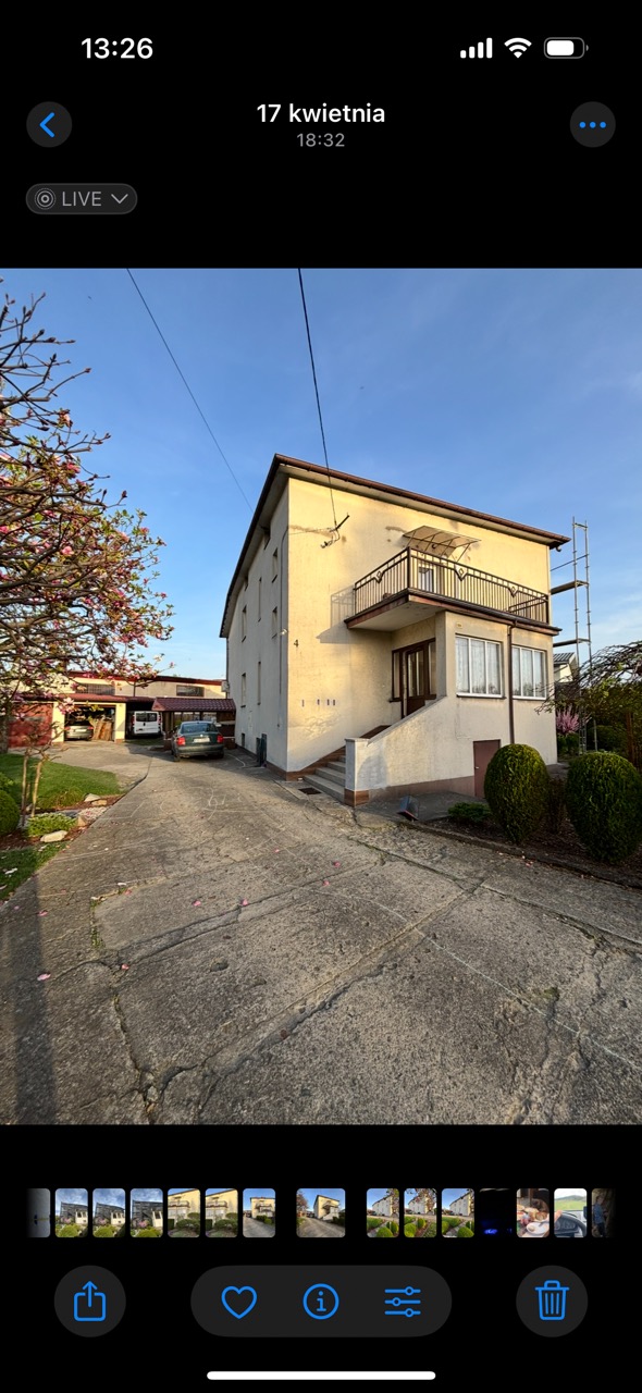 Dwupiętrowy dom z balkonem i podjazdem, widok z dołu. Na ścianie widoczne ślady po naprawach, obok rusztowanie. Wiosenne drzewo dodaje koloru.