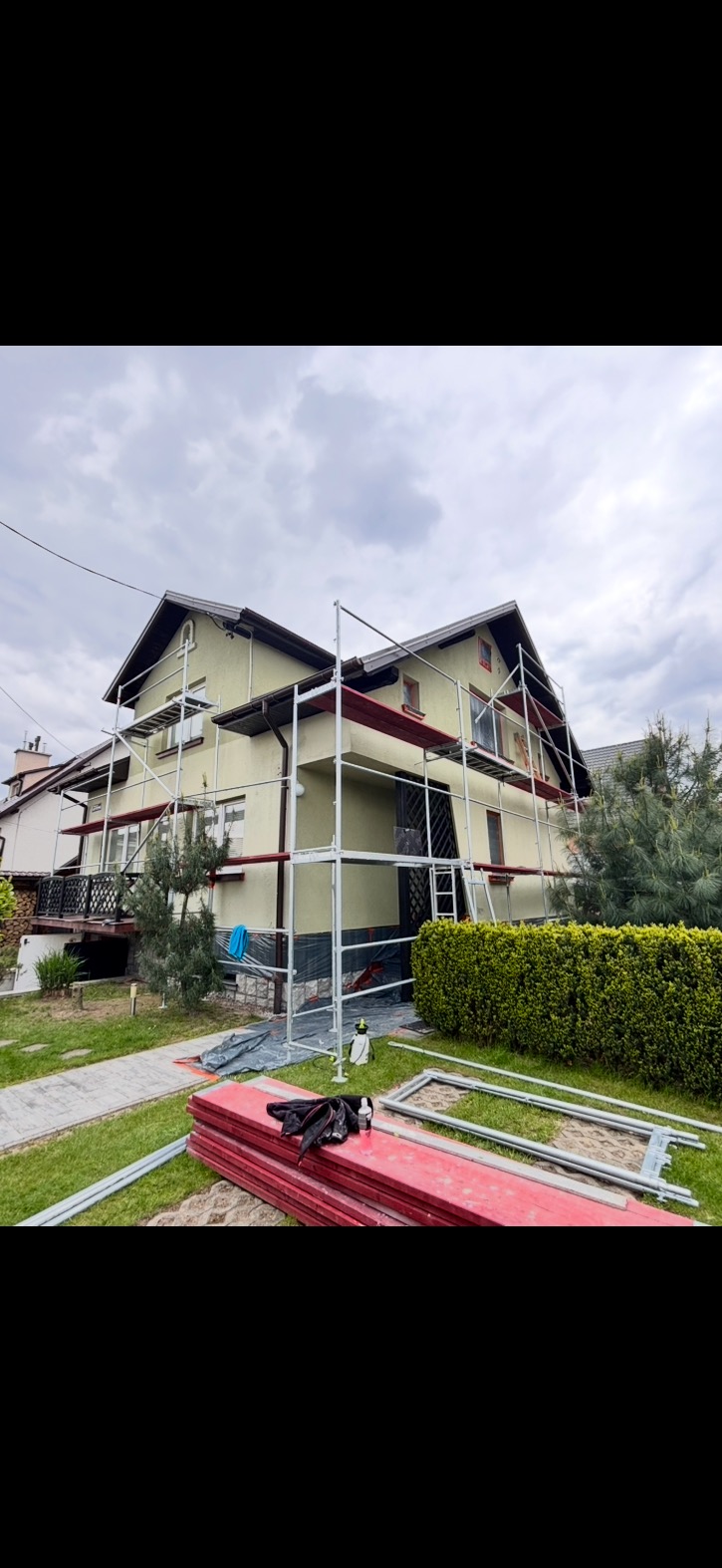Dom w rusztowaniach w Uściu Gorlickim, przygotowany do malowania elewacji. Widoczne materiały i narzędzia na trawniku, ściany w jasnym odcieniu.