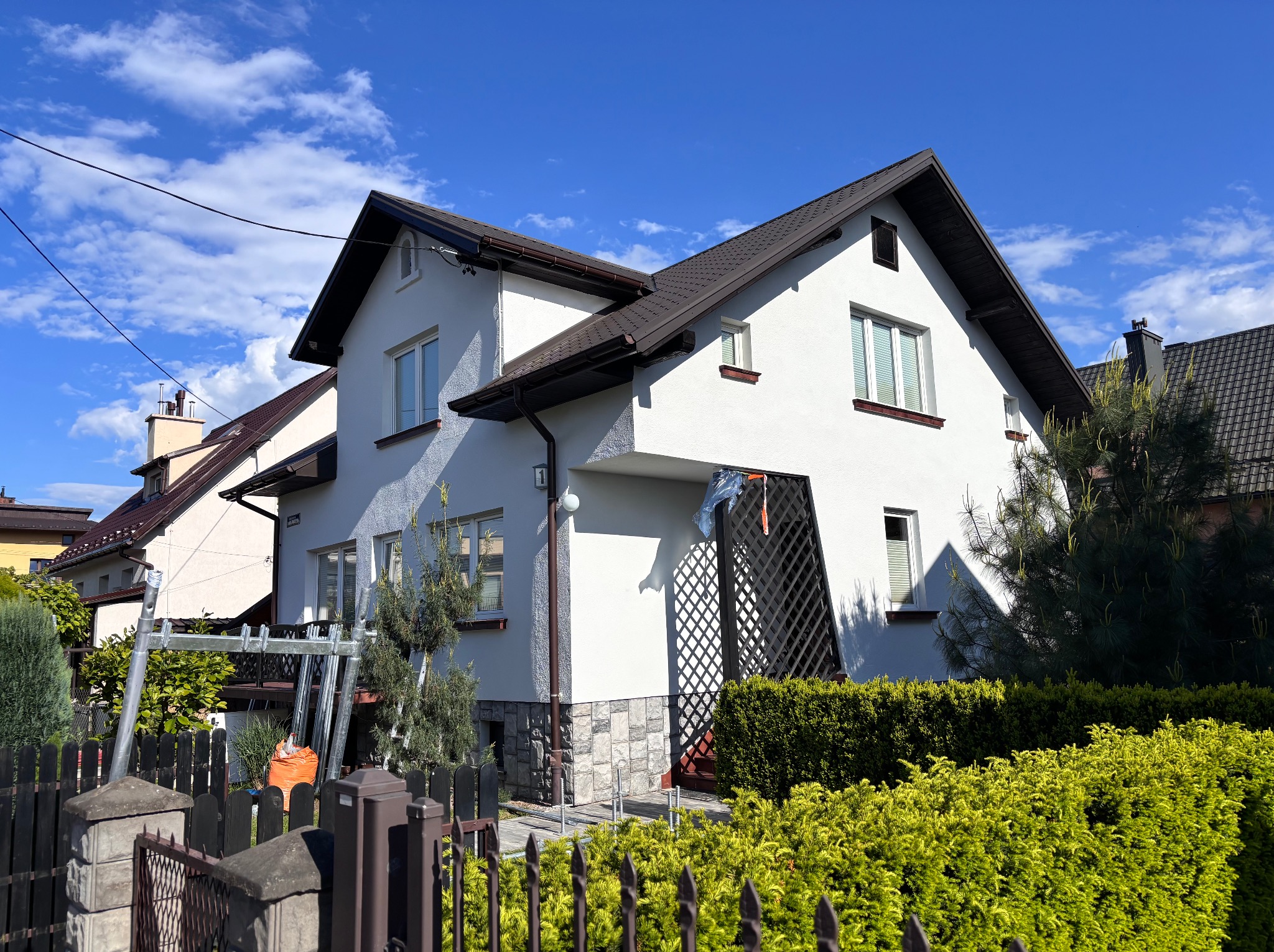 Dwupiętrowy dom z białą elewacją i brązowym dachem, widoczny zza ogrodzenia. Część elewacji pomalowana, obok domu rozstawione rusztowanie, w tle błękitne niebo.