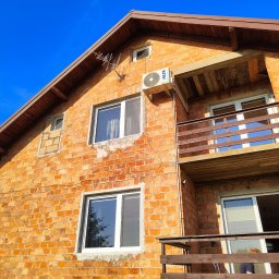 H20TRONIK Tomasz Dombrowski - Boczny widok na dwupiętrowy dom z czerwonej cegły z balkonem i zamontowaną jednostką klimatyzacji AUX na ścianie, antena telewizyjna na dachu, błękitne niebo w tle.