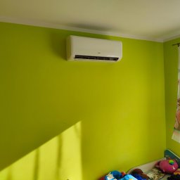 H20TRONIK Tomasz Dombrowski - Biały klimatyzator marki AUX zamontowany na limonkowo-zielonej ścianie w pokoju dziecięcym, poniżej widoczne łóżko z pościelą w bajkowe wzory i zasłonka z postaciami księżniczek.