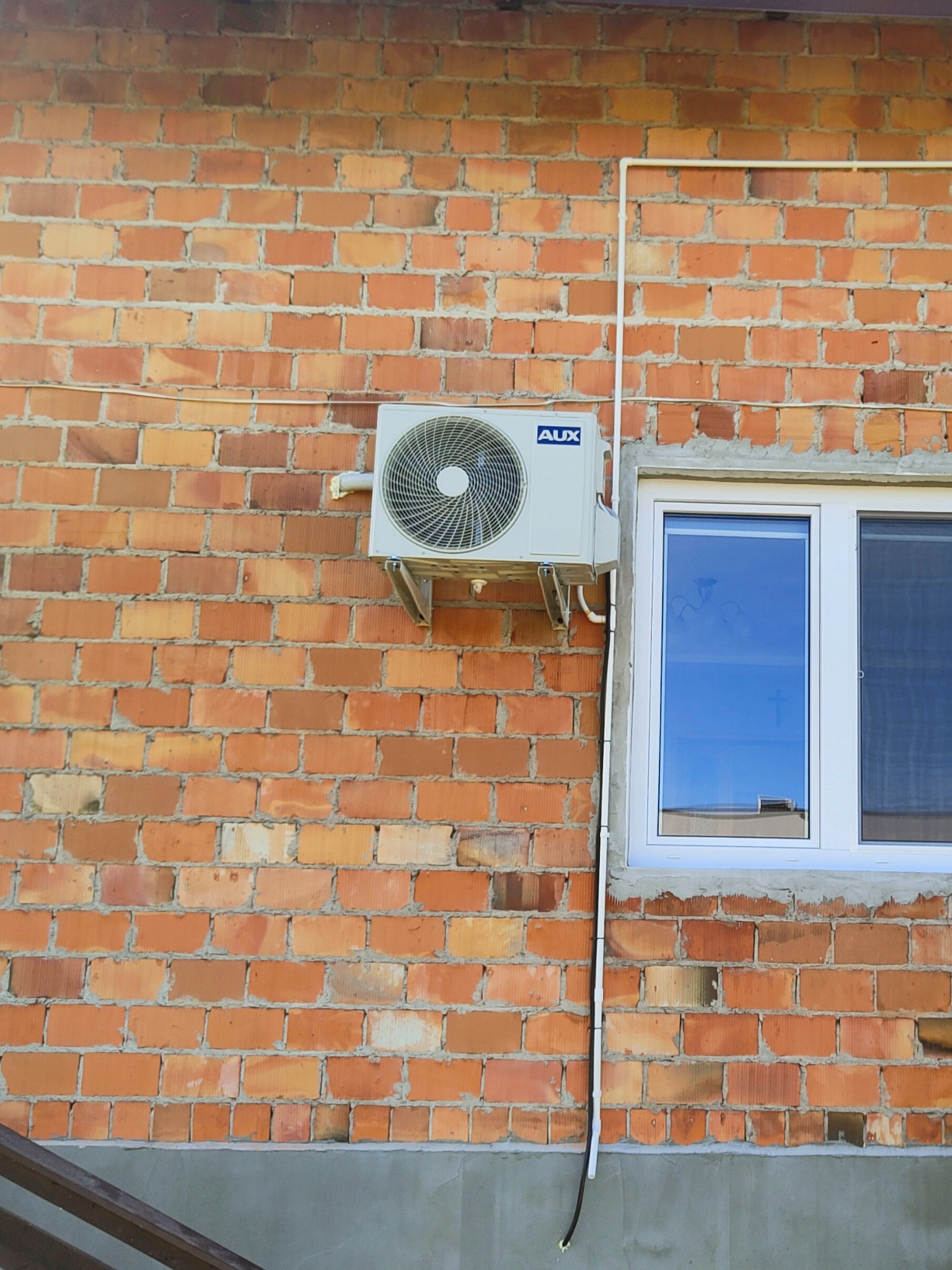 Zewnętrzna jednostka klimatyzacji AUX zamontowana na ceglanej ścianie budynku obok białego okna z widocznym odbiciem nieba. Kabel zasilający i rurka odprowadzająca skropliny poprowadzone wzdłuż...