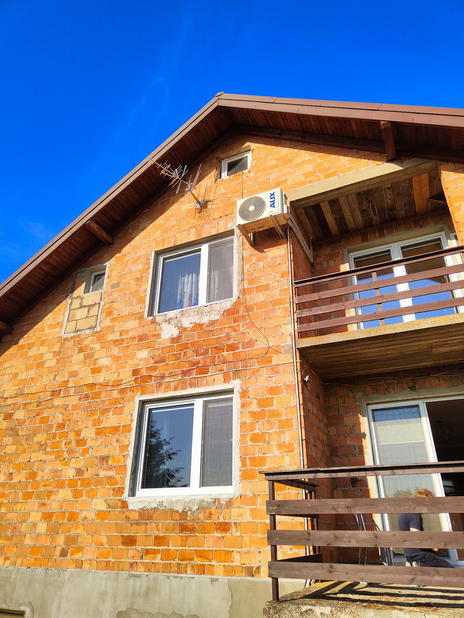 Boczny widok na dwupiętrowy dom z czerwonej cegły z balkonem i zamontowaną jednostką klimatyzacji AUX na ścianie, antena telewizyjna na dachu, błękitne niebo w tle.