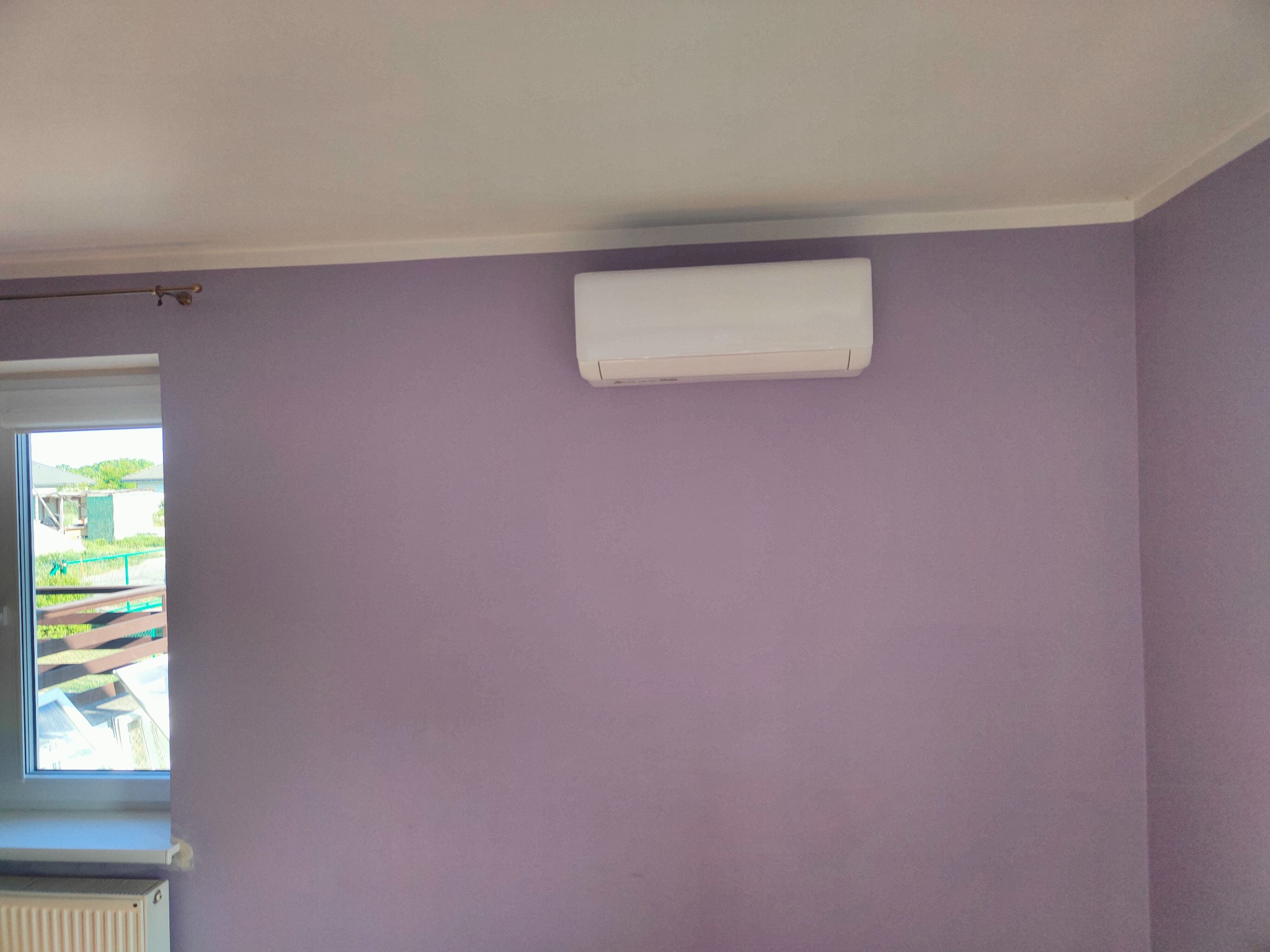 Biała jednostka klimatyzacji ściennej zamontowana w pokoju z fioletowymi ścianami, widoczne okno z widokiem na zewnątrz i grzejnik pod oknem.