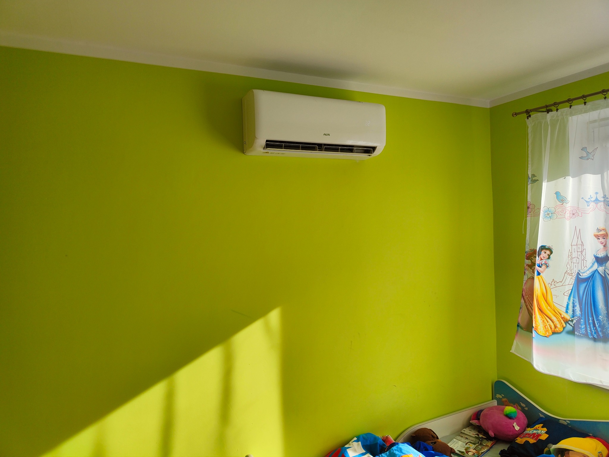 Biały klimatyzator marki AUX zamontowany na limonkowo-zielonej ścianie w pokoju dziecięcym, poniżej widoczne łóżko z pościelą w bajkowe wzory i zasłonka z postaciami księżniczek.