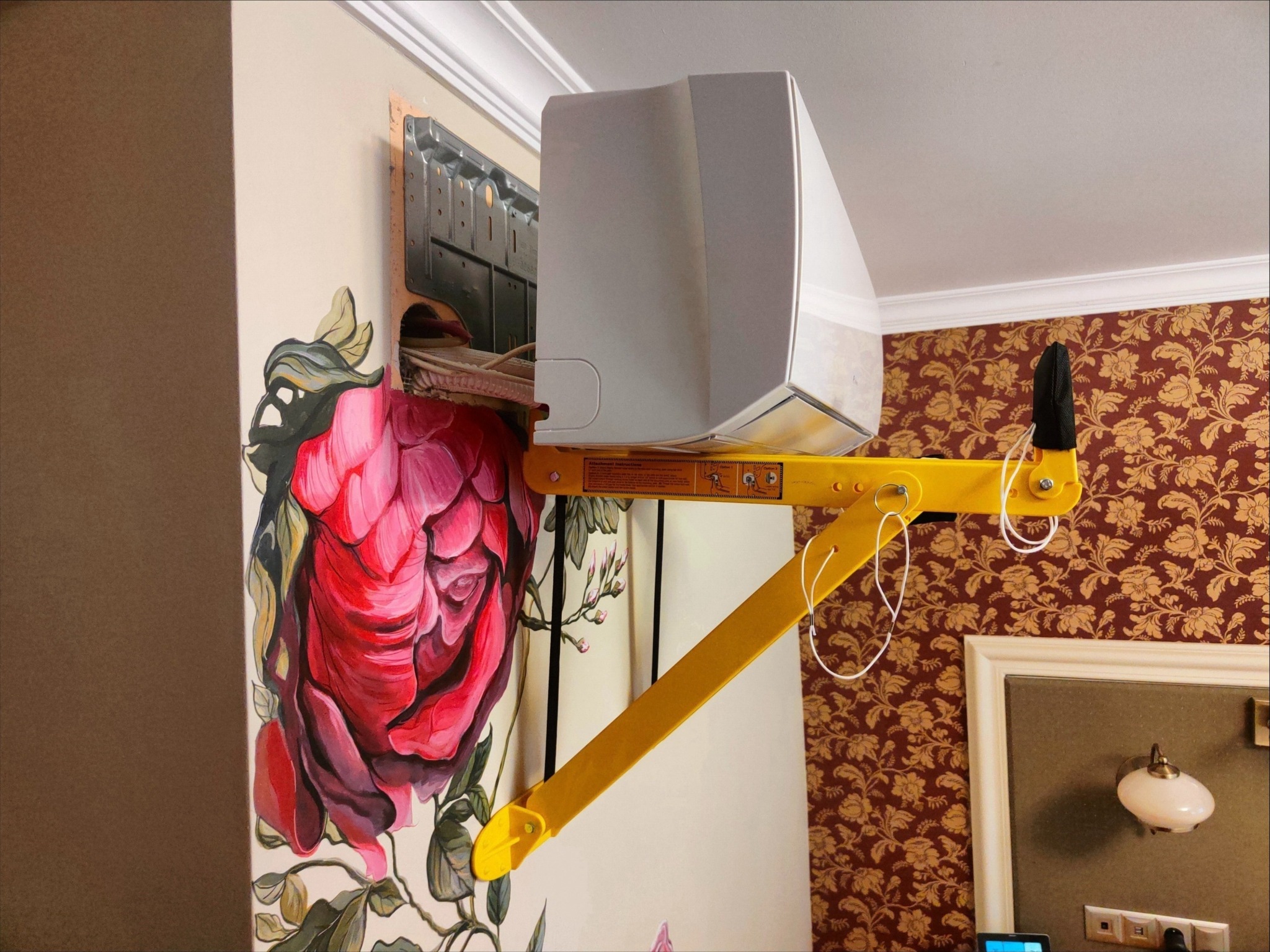 Montaż klimatyzatora na ścianie z kwiatowym motywem, widoczny żółty podnośnik, wnętrze z wzorzystą tapetą w tle.