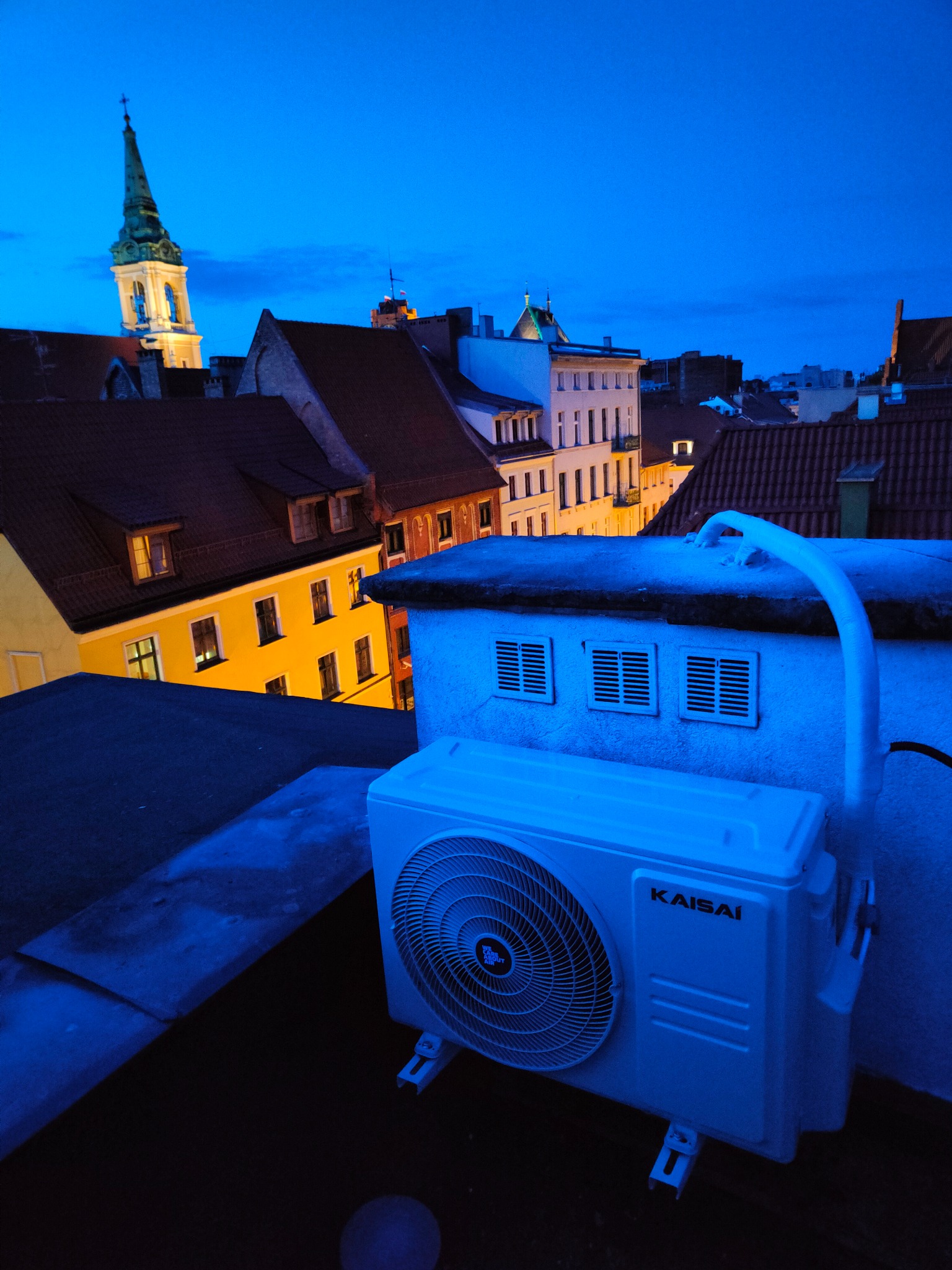 Jednostka zewnętrzna klimatyzacji Kaisai zamontowana na dachu budynku w Silno, widok na starówkę w niebieskiej poświacie zmierzchu.