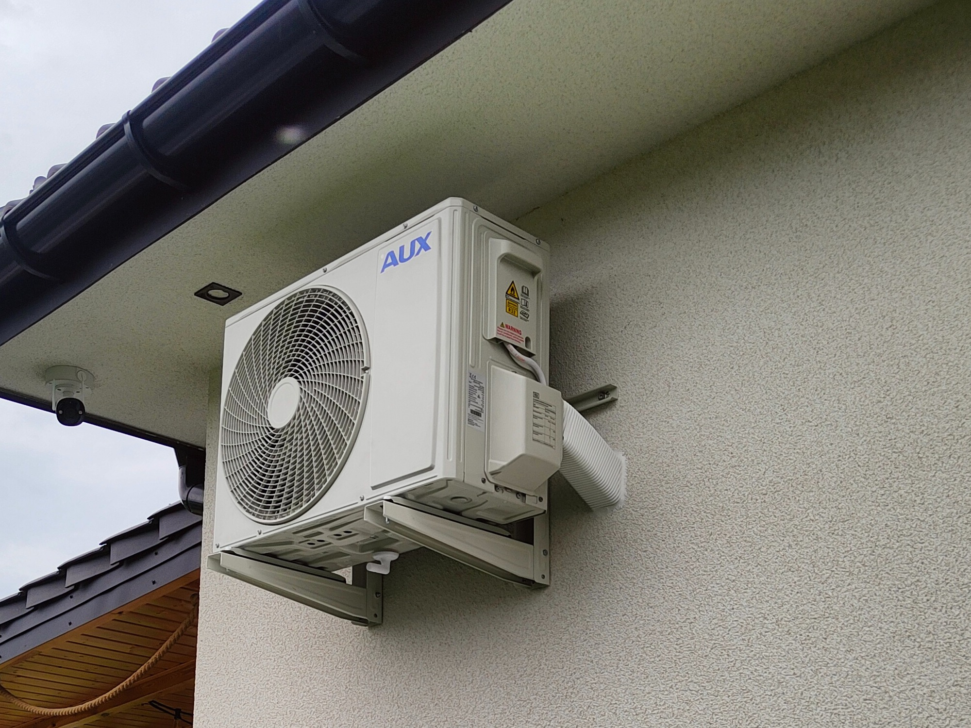 Zewnętrzna jednostka klimatyzacji marki AUX zamontowana na elewacji domu z fakturą baranka, widoczna kamera monitoringu pod dachem.