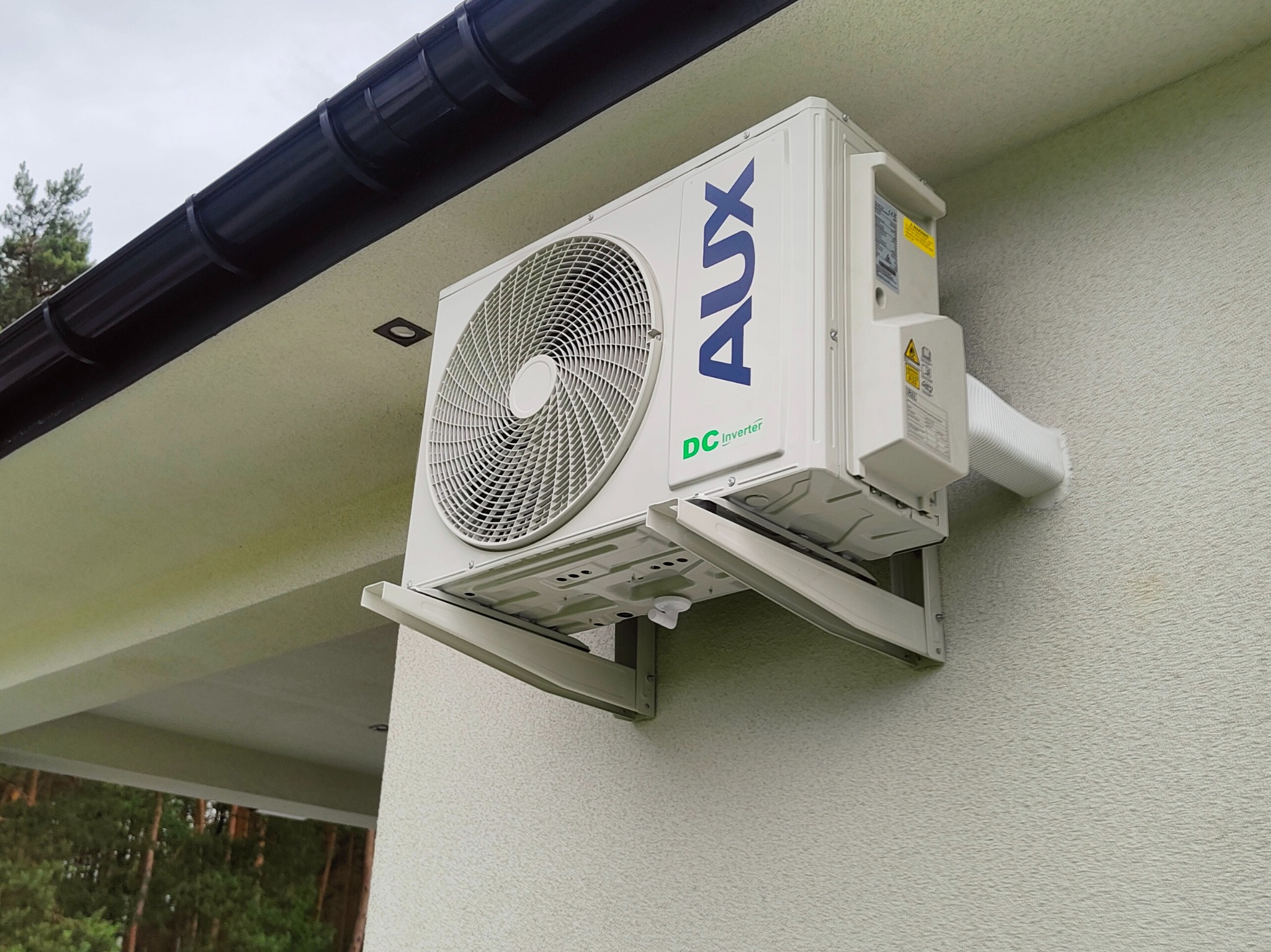 Zewnętrzna jednostka klimatyzacji AUX DC Inverter zamontowana na elewacji budynku, widoczna instalacja odprowadzająca skropliny, tło stanowią drzewa.