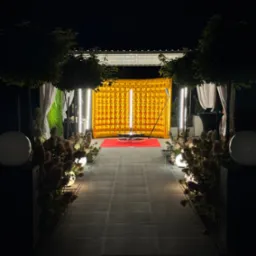 Oświetlona fotobudka z dmuchanym złotym tłem, ustawiona na czerwonym dywanie na zewnątrz, otoczona kwiatami i kulami świetlnymi na tle nocnego nieba.