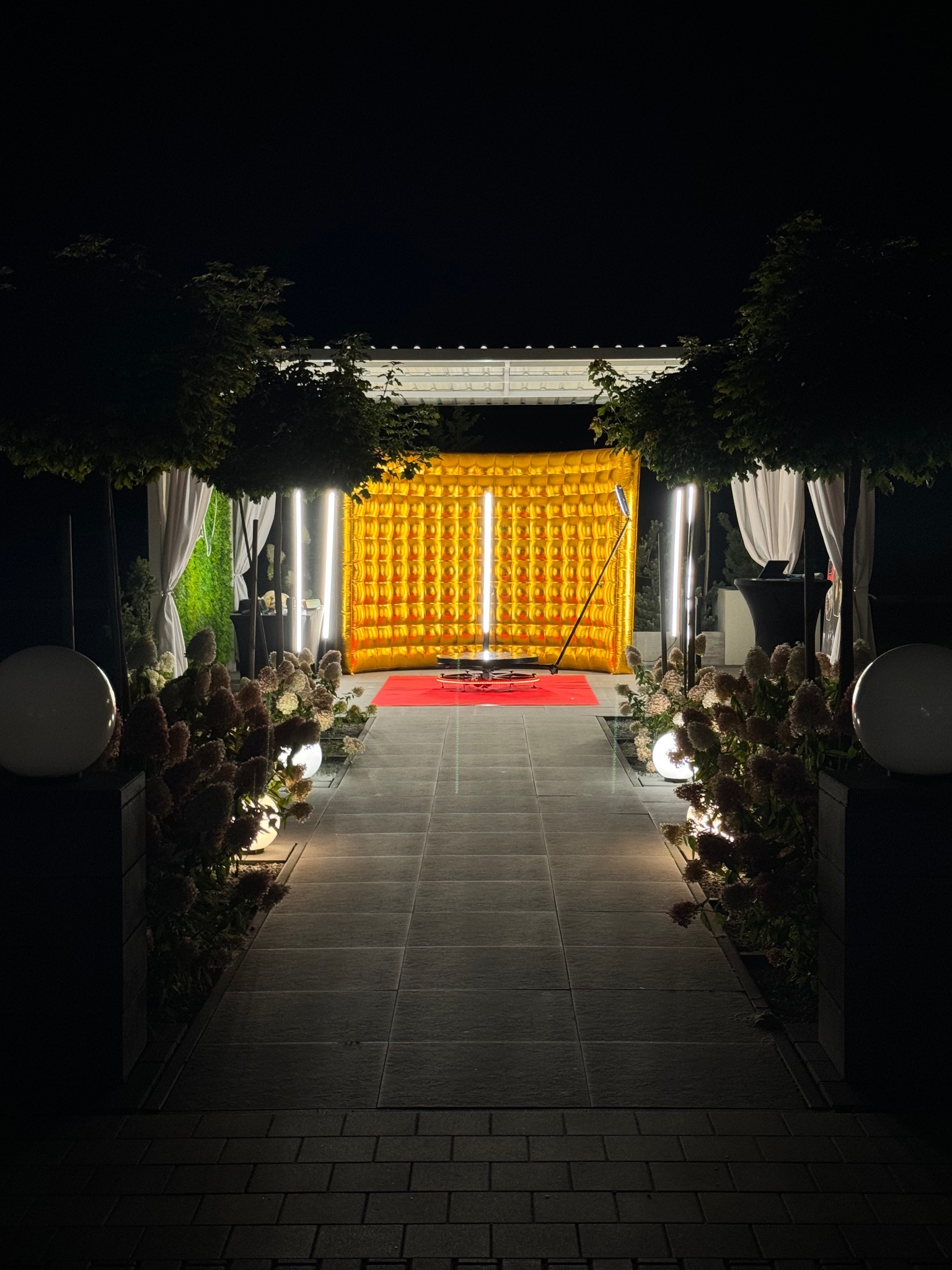 Oświetlona fotobudka z dmuchanym złotym tłem, ustawiona na czerwonym dywanie na zewnątrz, otoczona kwiatami i kulami świetlnymi na tle nocnego nieba.