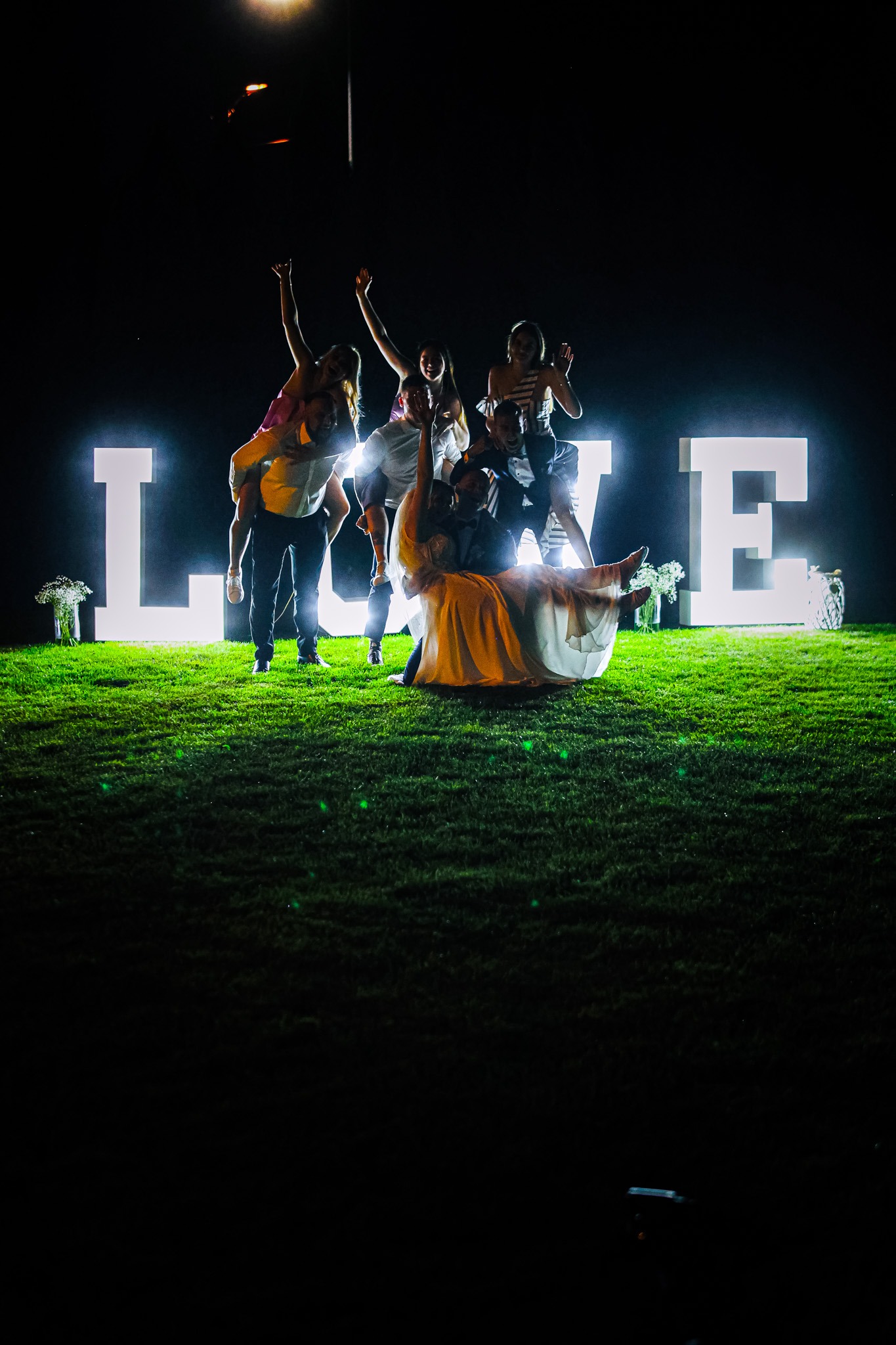 Grupa osób pozuje na trawie przed dużymi, podświetlanymi literami 'LOVE' podczas nocnej imprezy.